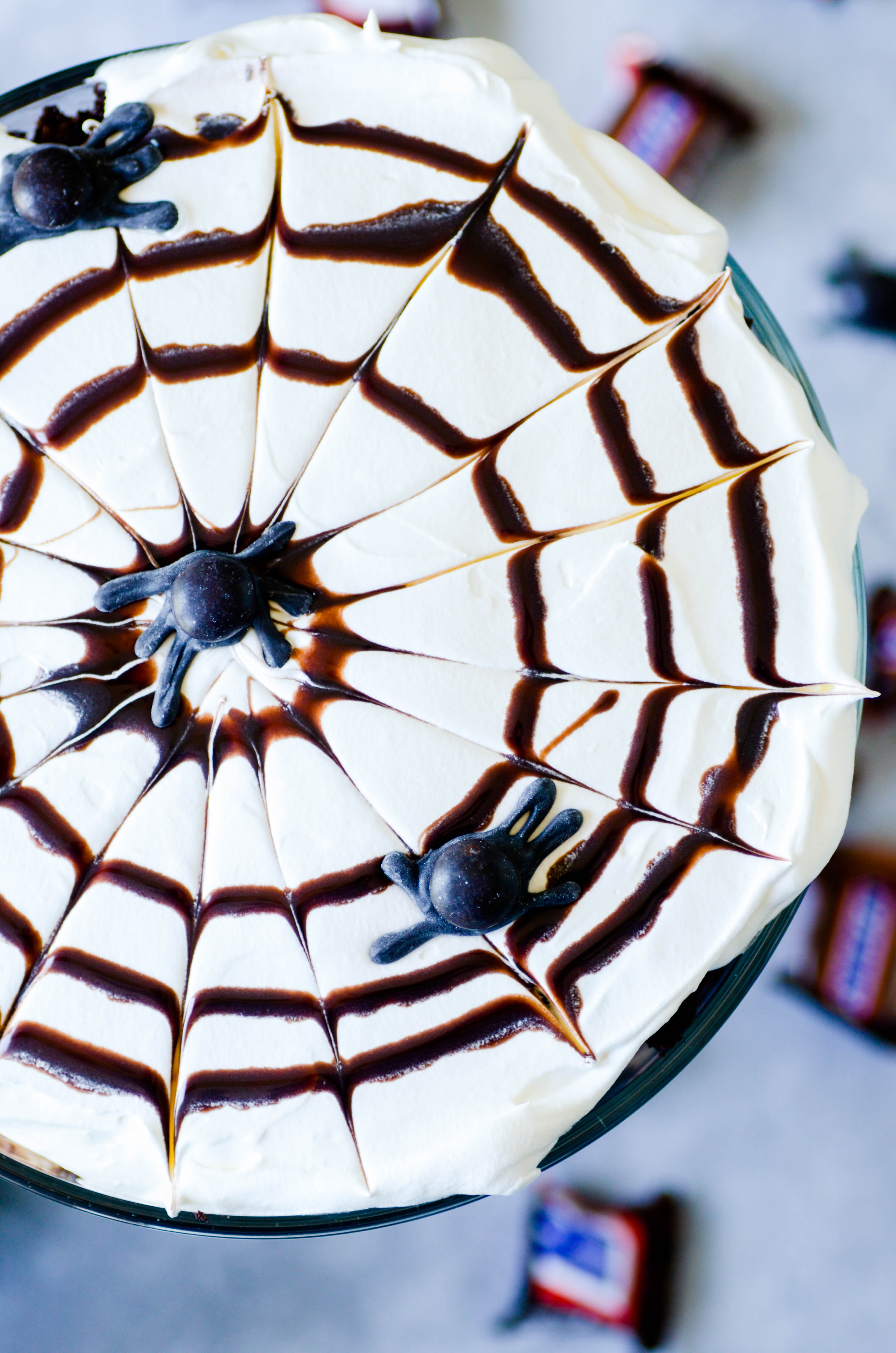 Layers of cake, pudding, whipped cream, and snickers come together in a simple and delicious dessert topped with a chocolate syrup spider web.